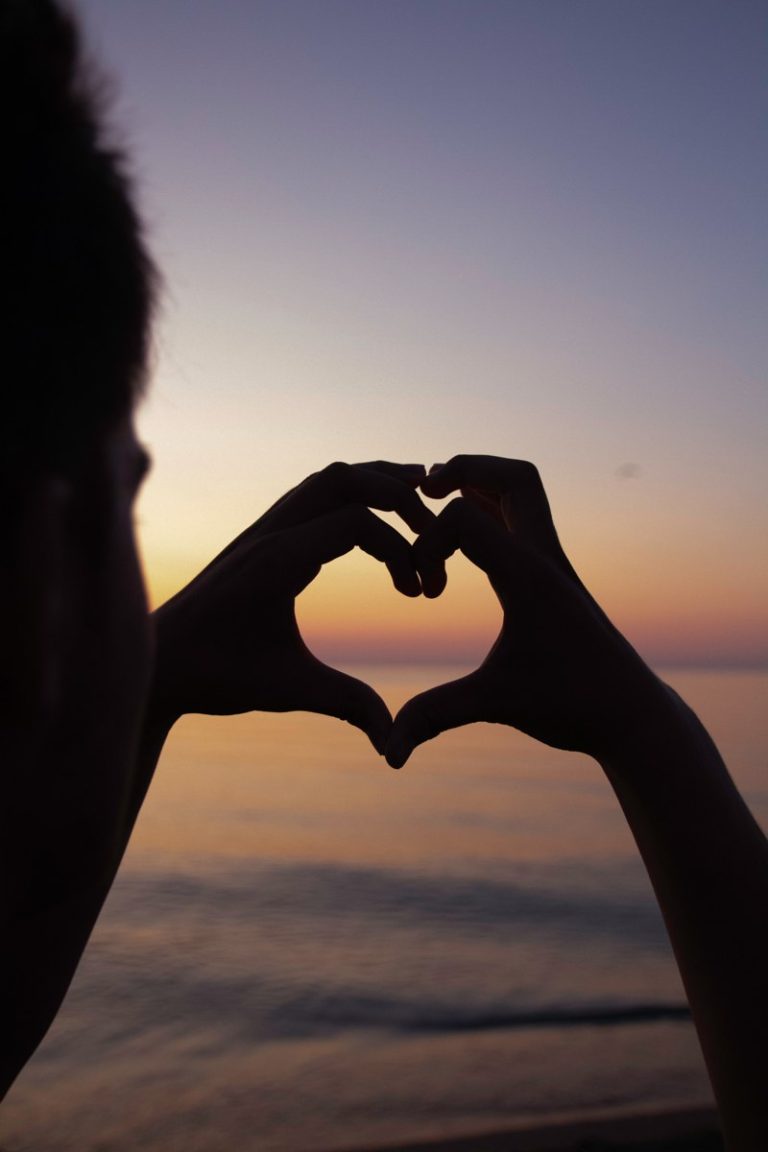 Hands making a heart shape against a sunset over the water